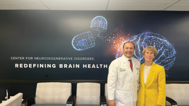 a doctor in a white coat and a woman in a yellow blazer stand in front of a sign for the Center for Neurogenerative Disorders